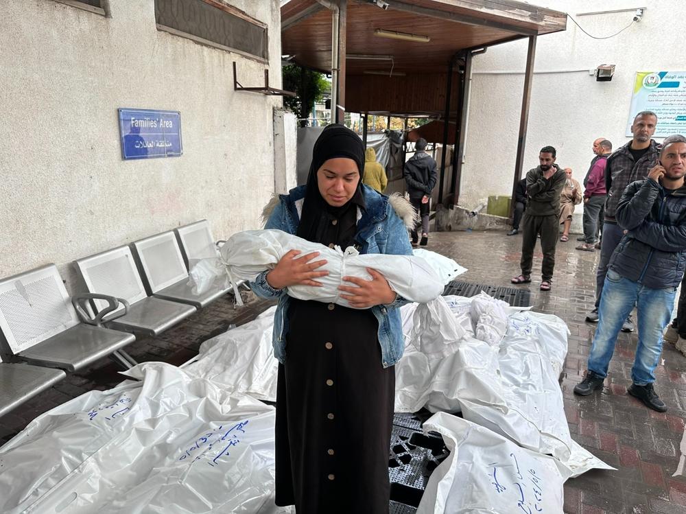 A woman carries a child in a white shroud in front of a 