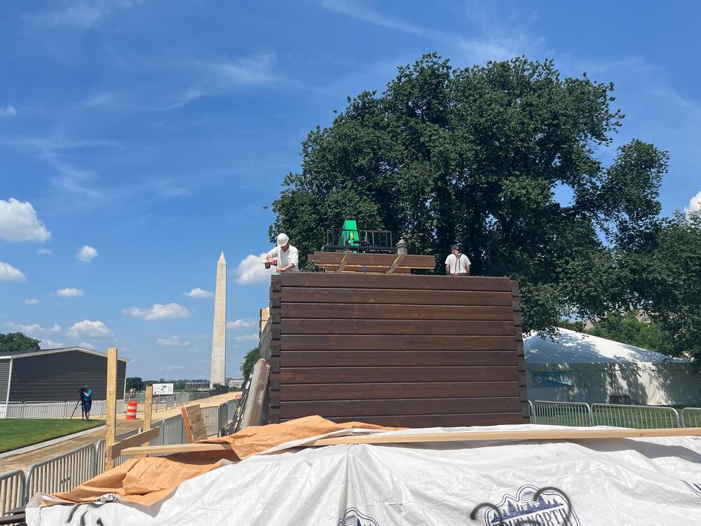 Workers assemble a home resembling an instant log cabin on the National Mall in Washington, D.C., with the Washington Monument behind them.