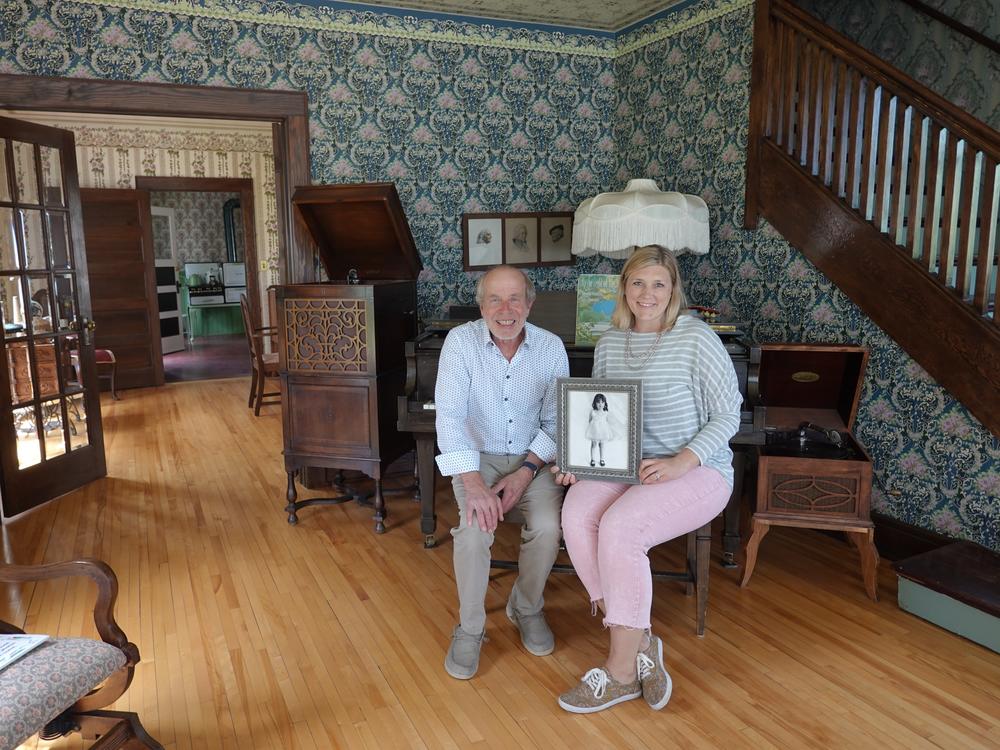 Two people sitting with photo: John Kelsch, left, and Janie Heitz, right, holding a framed photo of a young Judy Garland. They are the former and current directors of the Judy Garland Museum in Grand Rapids, Minnesota. They posed for a photo in the actress’ childhood home on May 30.