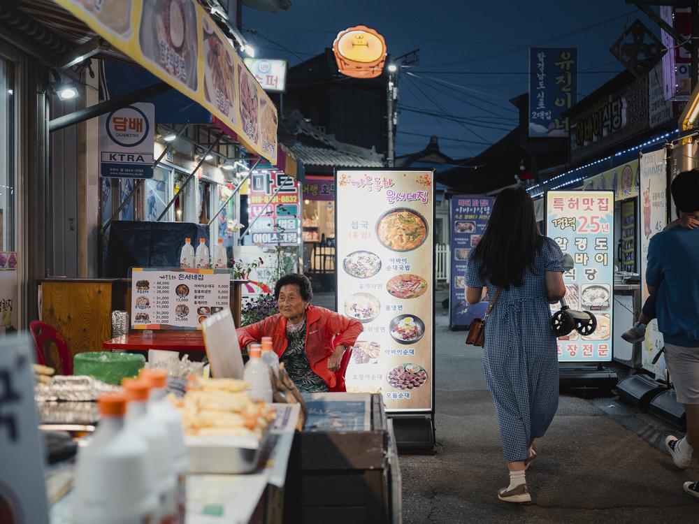 Jun Sook-ja, 91 (left), sits at a restaurant in Abai Village as tourists walk by on Sept. 9, 2023. The village has become a tourist destination known for being a village of displaced people who settled here after the Korean War.
