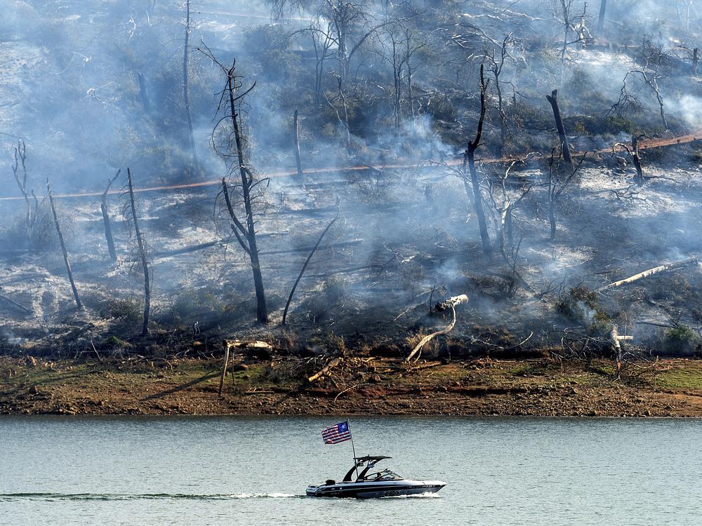 A boat crosses Lake Oroville with a smoldering hillside behind as the Thompson Fire burns in Oroville, Calif., on Wednesday. An extended heatwave blanketing Northern California has resulted in red flag fire warnings and power shutoffs.