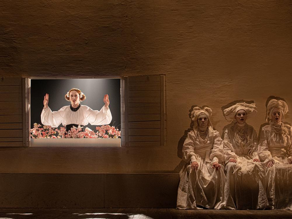 A scene from a Ukrainian production of <em>The Witch of Konotop.</em> At the window, Olena, played by Mariia Rudynska, is the love interest of the main character but does not love him back. And a three-witch chorus is seen right.