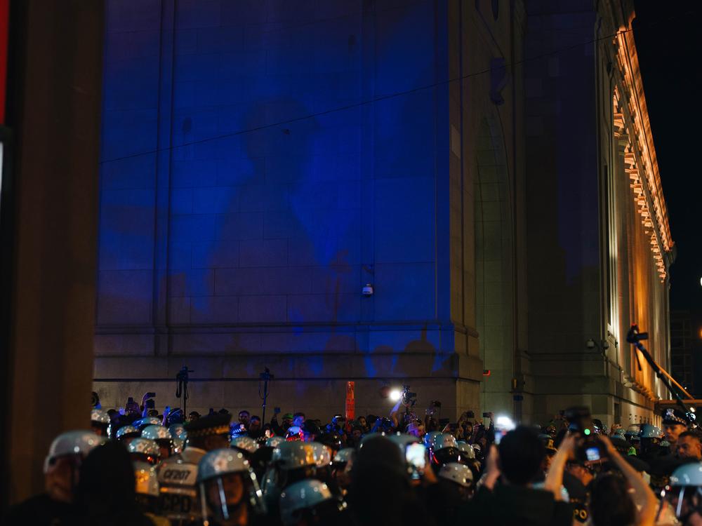 The shadow of a police officer shines against a wall as protestors and officers crowd below.