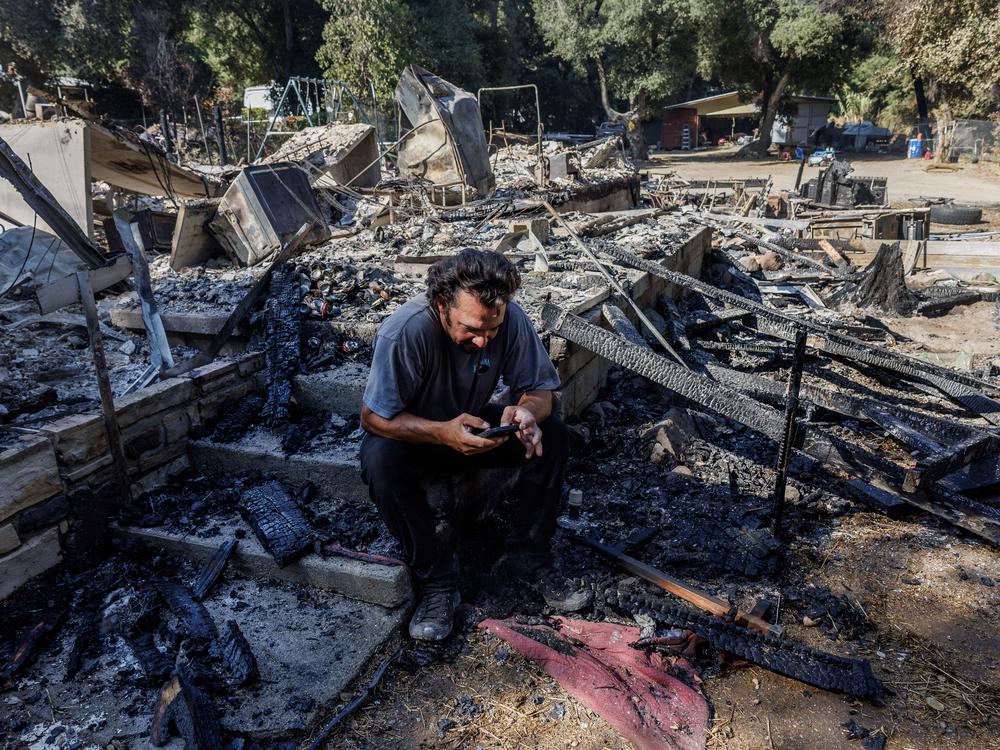Garrett Keene sits on the front steps of his home in Lake Elsinore, Calif. that was destroyed in the Airport Fire, one of three major Southern California Fires so far this September. After evacuating with his family, Keene said he monitored the Watch Duty fire app to see where airdrops of fire retardant were happening as the fire approached, but none dropped on his neighborhood.