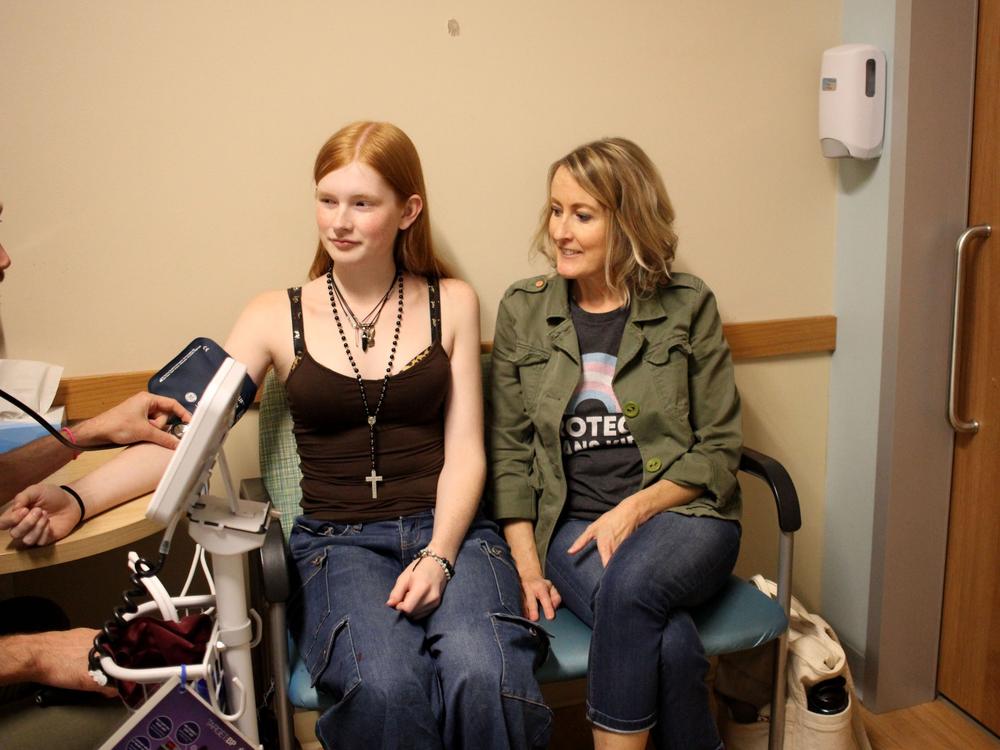Veronica’s Minneapolis pediatrician takes her blood pressure. During the appointment, Veronica receives a puberty blocker injection and a refill of her prescription estrogen pills. Emily is wearing a t-shirt that says 