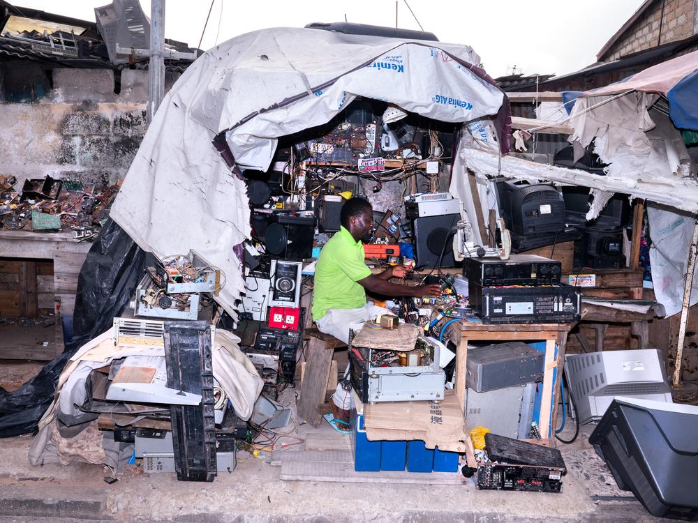 The man in the bright green shirt is working in one of the <strong>h</strong>undreds of small shops that have popped up around e-waste sites, where discarded items are repaired and resold, or broken down for parts.
