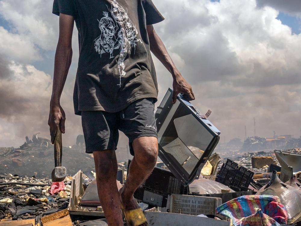 An 18-year-old holding a hammer and a TV walks over pile of e-waste. Collecting e-waste to salvage can be dangerous work. Cuts, burns and other injuries are common, and e-waste sites are often blanketed in noxious fumes.