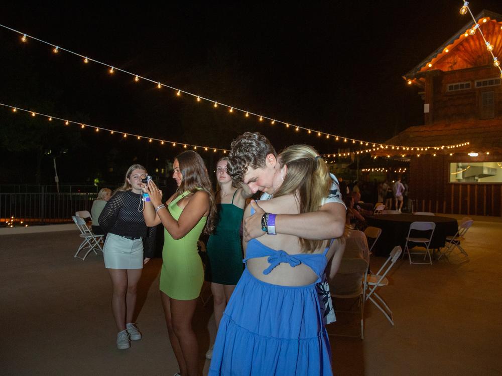 A young couple embraces while their friends take selfies and pictures on the deck underneath the stars.