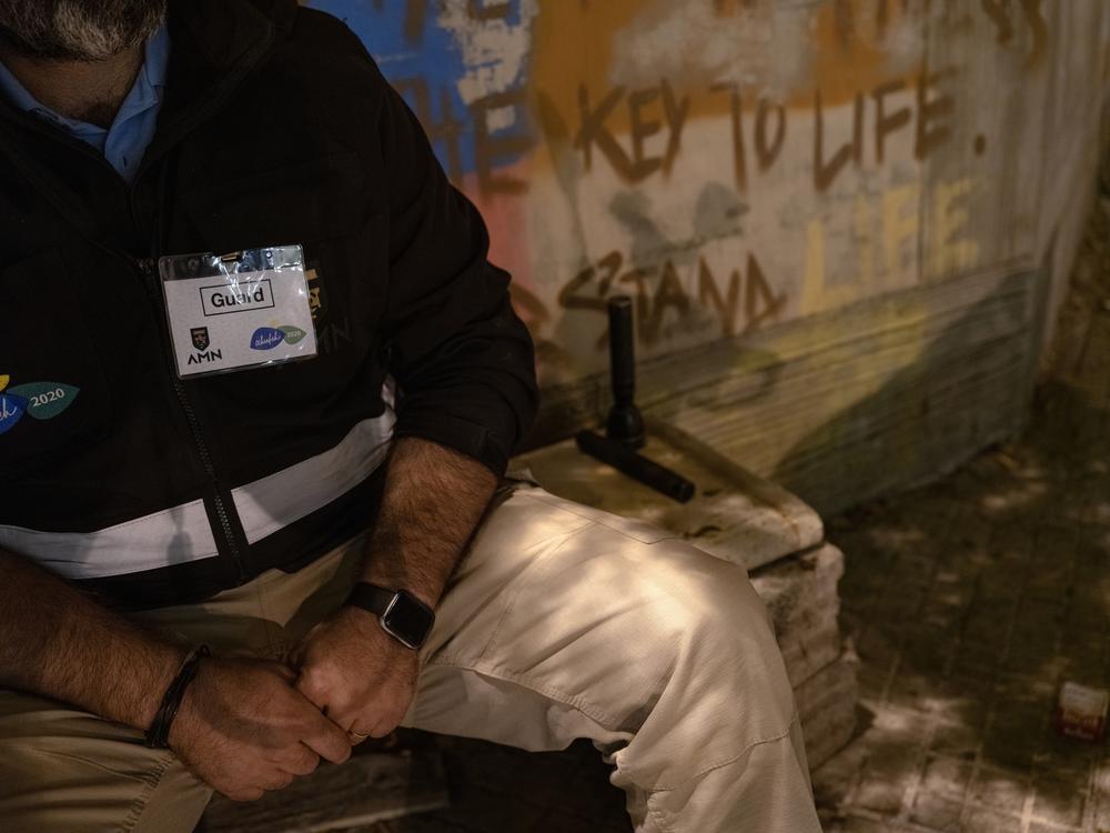 A night guard in Ashrafieh hides in plain sight, in a dark street corner, on high alert for any suspicious individuals.