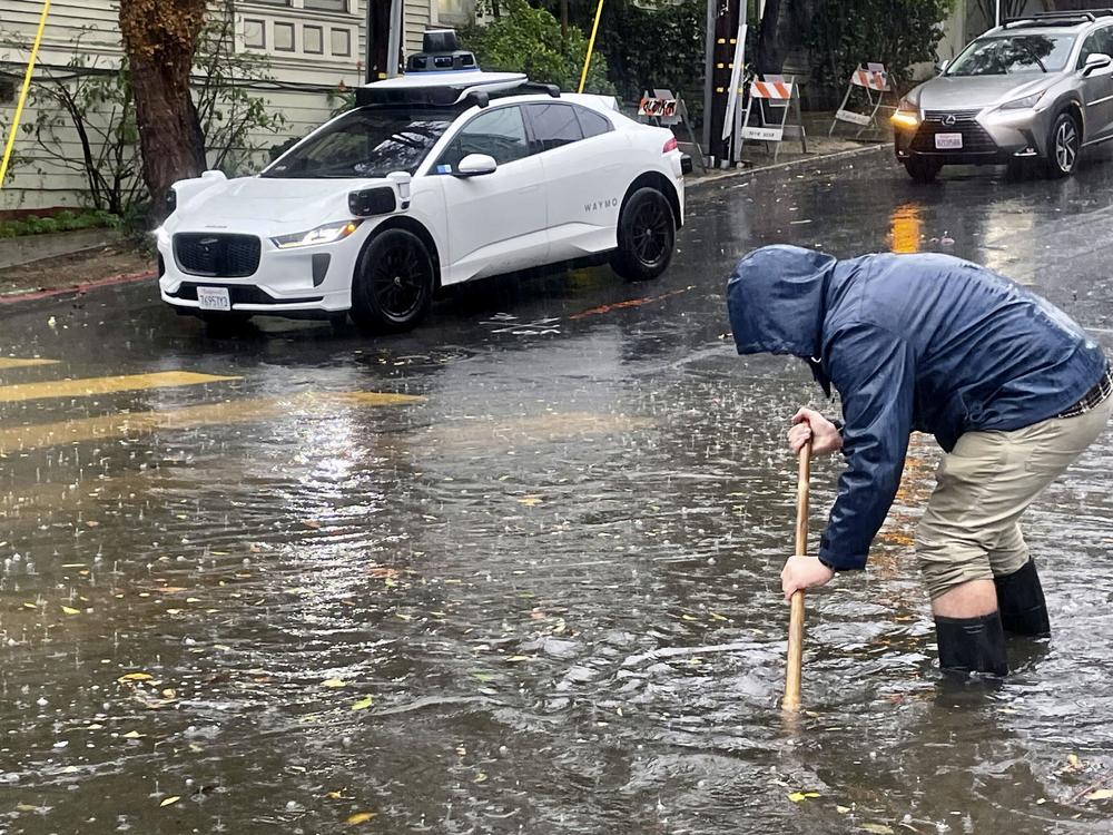 A Mission District resident attempts to unclog a few storm drains on a flooded street as a driverless Waymo taxi passes in the background, Friday in San Francisco.