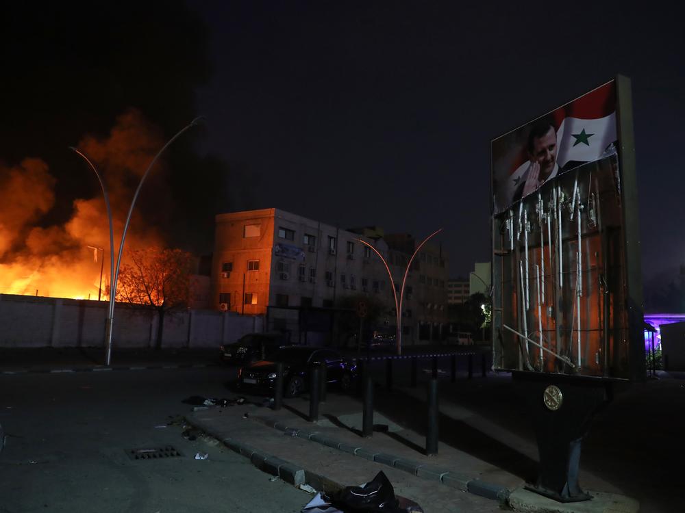 A damaged poster of Bashar al-Assad is seen as a fire burns at a security compound that houses the Syrian Military Intelligence Interrogation Division, in Damascus, Syria.