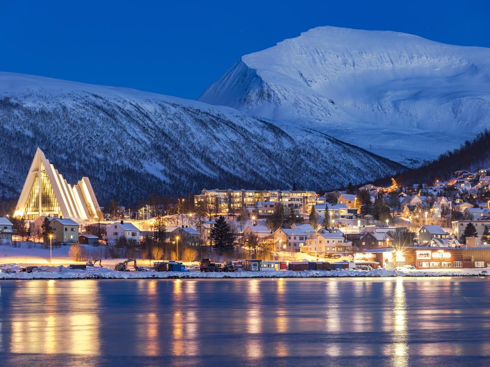 Dusk falls over snowy mountains that overlook the Arctic Cathedral and the houses mirrored in the icy fjord in Tromsø, Norway. Although this city is north of the Arctic Circle and experiences polar night for two months out of the year, its residents tend to see winter as a time of opportunity.