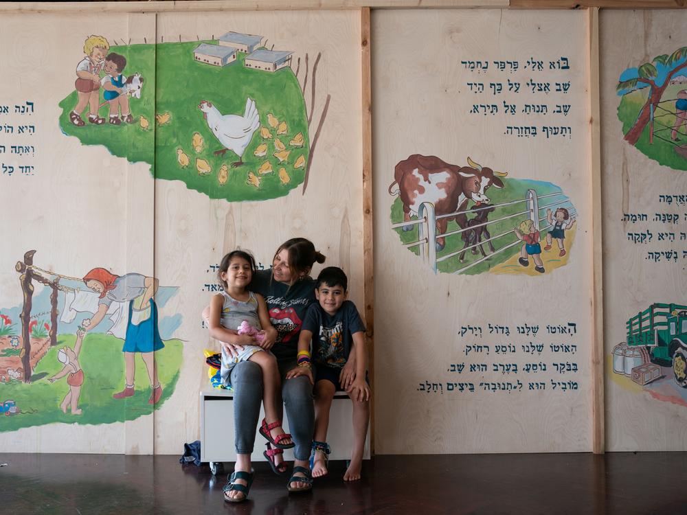 Alice Shahar, 42, poses with her children, Achinoam, 3, and Tzabar, 6, in front of a mural depicting a classic Israeli children's book's pages, originally printed in the Be'eri printing press, the kibbutz's main source of income. Shahar was the coordinator of seven kindergarten classes the displaced kibbutz residents established at the hotel for the Be'eri community. The residents brought the mural with them to stay connected to their home.