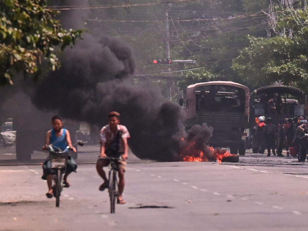 Cyclists pass police standing next to a burning makeshift tyre barricade, erected by protesters demonstrating against the military coup, in Yangon's South Okkalapa township on April 1, 2021.