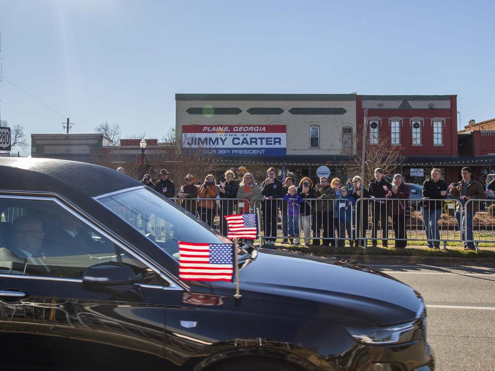 People watch as the hearse carrying the flag-draped casket of former President Jimmy Carter departs Phoebe Sumter Medical Center.