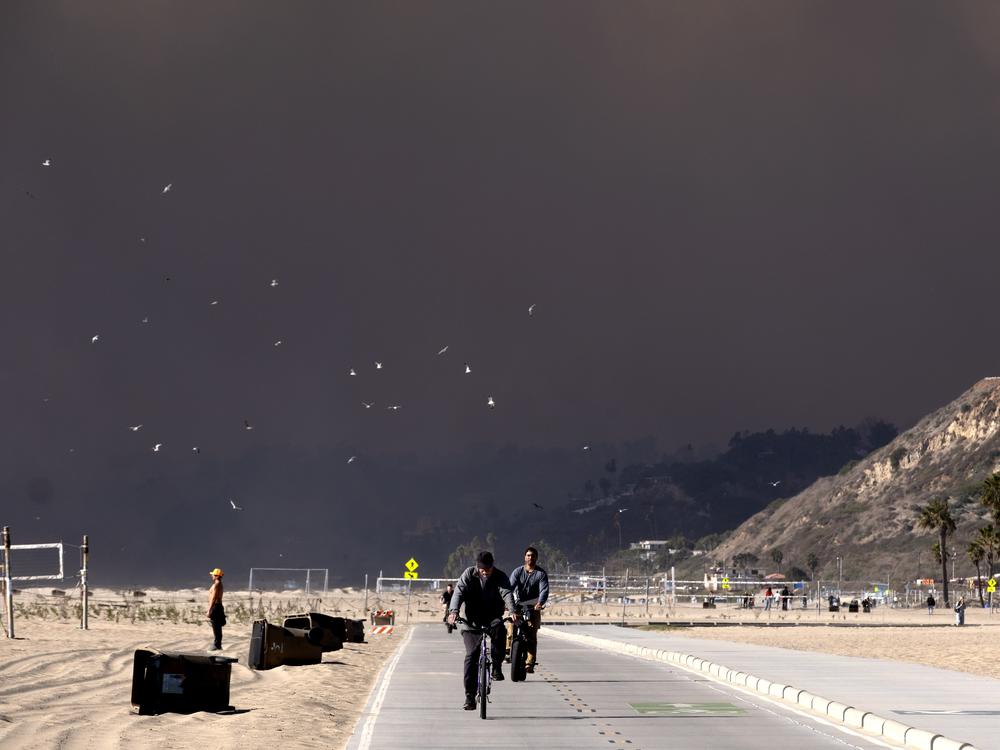 Bike riders make their way along the coast as large dark plume of smoke passes over the beach from a wildfire from Pacific Palisades, from Santa Monica, Calif.