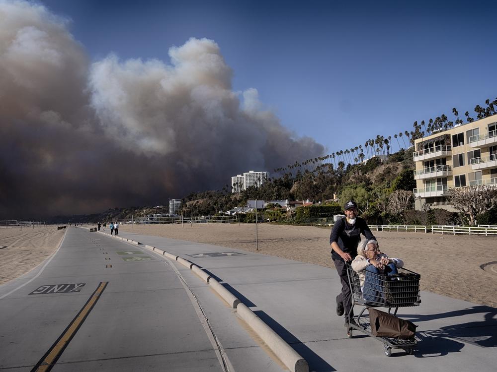 Jerome Krausse pushes his mother-in-law in a shopping cart as they evacuate from their home in the Pacific Palisades after a wildfire swept through their neighborhood in Santa Monica, Calif.