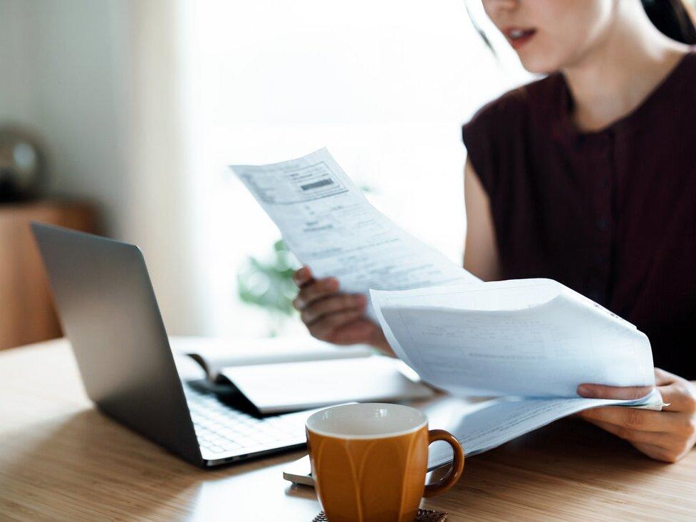 Cropped shot of Asian woman sitting at dining table, handling personal finance with laptop. She is making financial plan and planning budget as she go through her financial bills, tax and expenses at home. Wealth management, banking and finance concept
