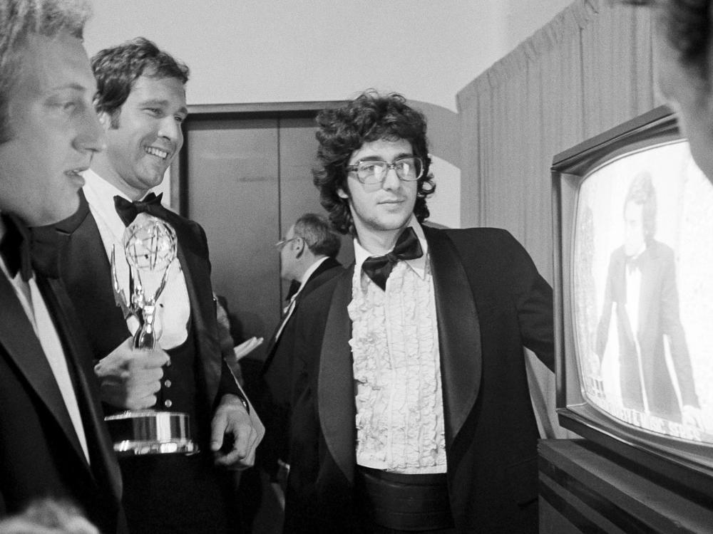 Chevy Chase, center, holds his Emmy Award as he watches producer Lorne Michaels accepting another Emmy for <em>SNL</em>, while writers Alan Zweibel, left, and Tom Davis look on in 1976.