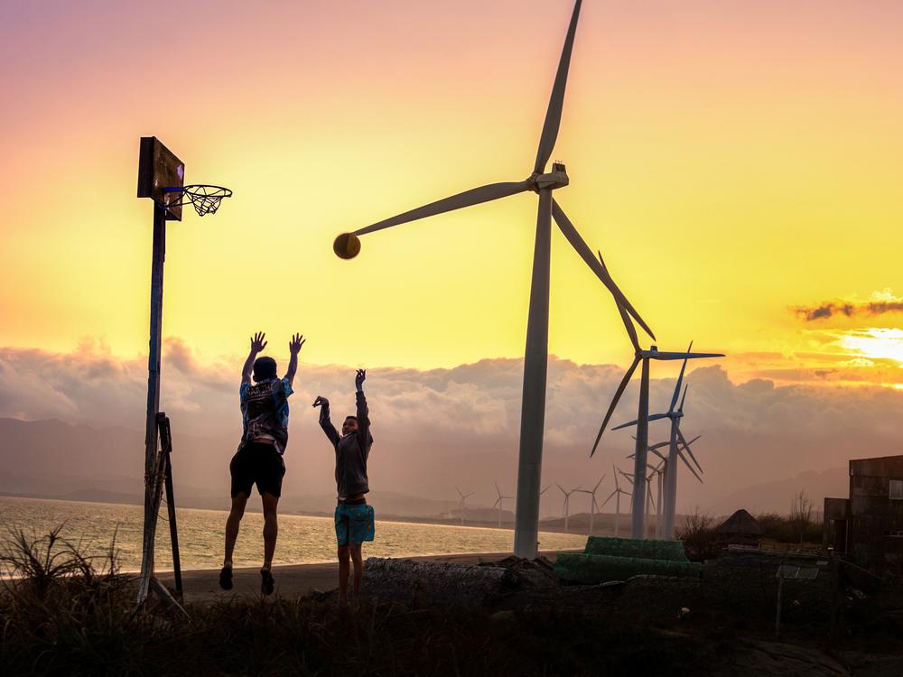 Two young people play basketball under the towering blades of the wind turbines in Bangui. They lie along a 5-mile shoreline of Bangui Bay, facing the South China Sea. Renewable energy has transformed this community, cutting household expenses and powering opportunities once thought to be out of reach.