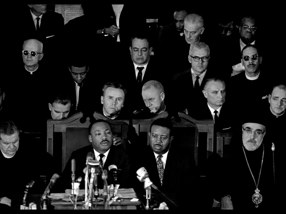 Dr. Martin Luther King, Jr. (center left), Rev. Ralph Abernathy (center right), and Archbishop Iakovos (far right) attend a memorial service for James Reeb. Reeb, a white Unitarian minister from Massachusetts who had answered Dr. King's call for religious leaders to join the protests, was brutally attacked as he left a local Black-owned restaurant on March 9. He died two days later.