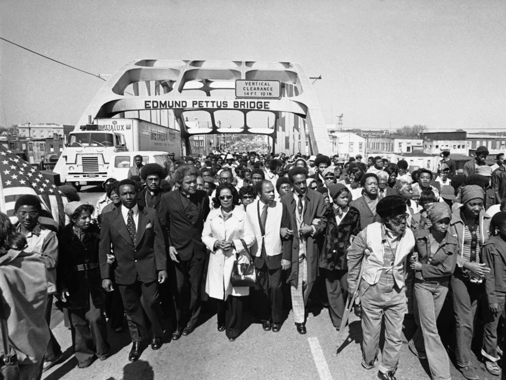 Aided by Father James Robinson, Mrs. Coretta Scott King, widow of Dr. Martin Luther King, Jr., center, and John Lewis, a crowd estimated by police at 5,000, marched across the Edmund Pettus Bridge from Selma, Ala. March 8, 1975.