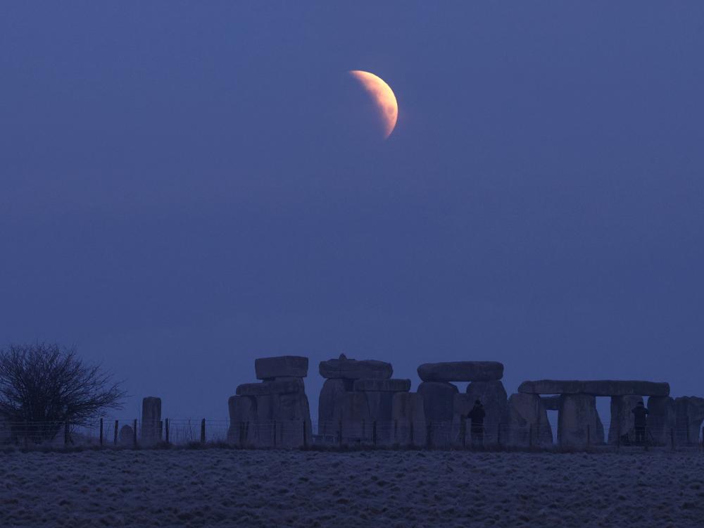 People view the full moon as a section moves into shadow during a lunar eclipse, before dawn on Friday, at Stonehenge stone circle, near Amesbury, southern Britain.