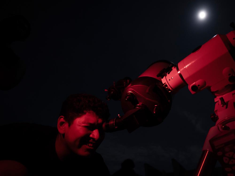 A man looks through a telescope at the full moon, also known as the 