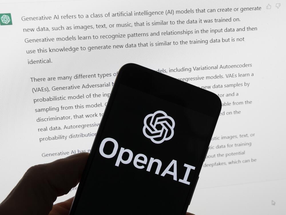 OpenAI's logo is on the screen of a mobile phone that's being held in front of a computer screen displaying output from ChatGPT.
