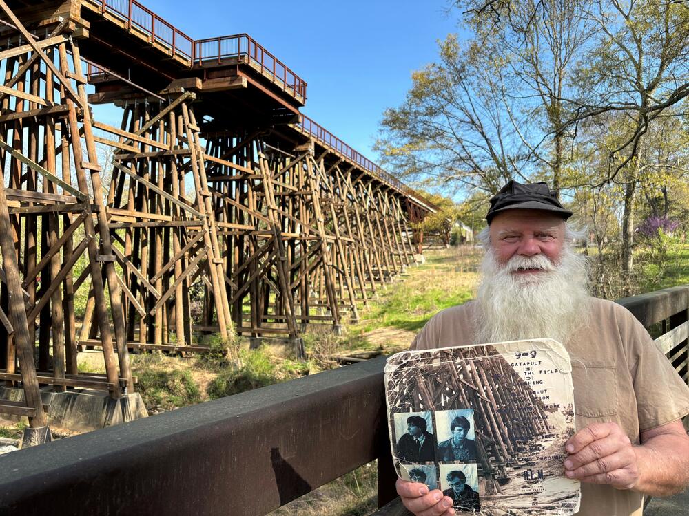 Paul Butchart gives tours of musical landmarks all over Athens, Ga. In his hands is R.E.M.'s 1983 <em>Murmur</em> album, with the original trestle bridge photograph on the back cover. The bridge behind Butchart is a reconstruction, and now part of a walking and biking trail.