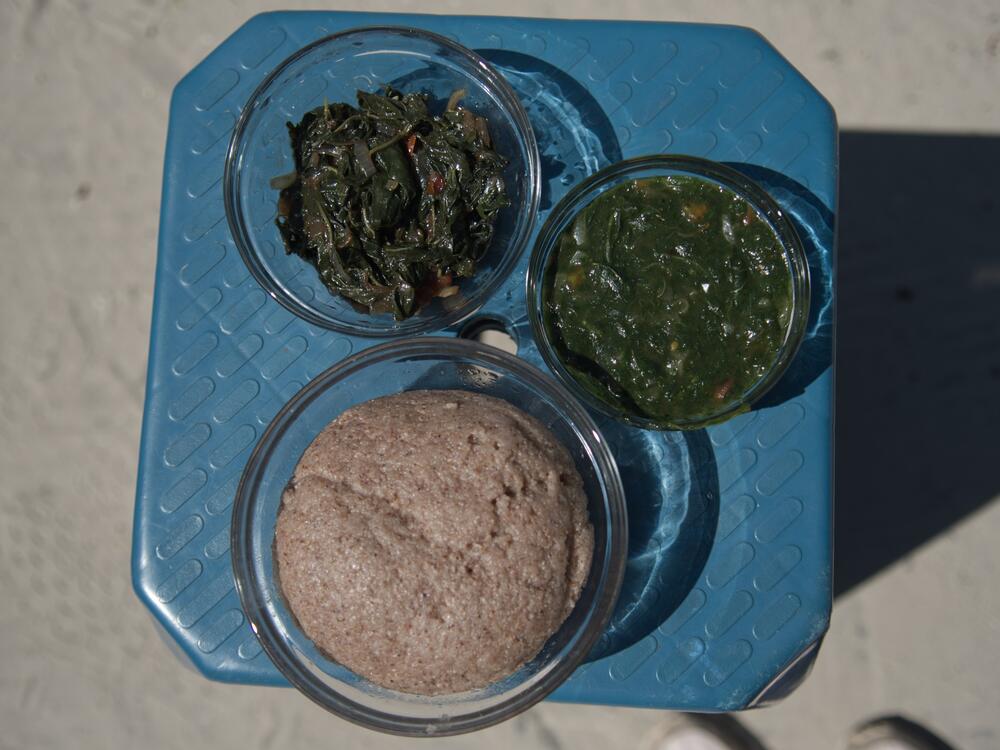 Two bowls of soup made with different types of local green leafy vegetables sit behind a bowl holding the grain sorghum, served as an accompaniment. The bowl on the right shows the soup in the recipe below.