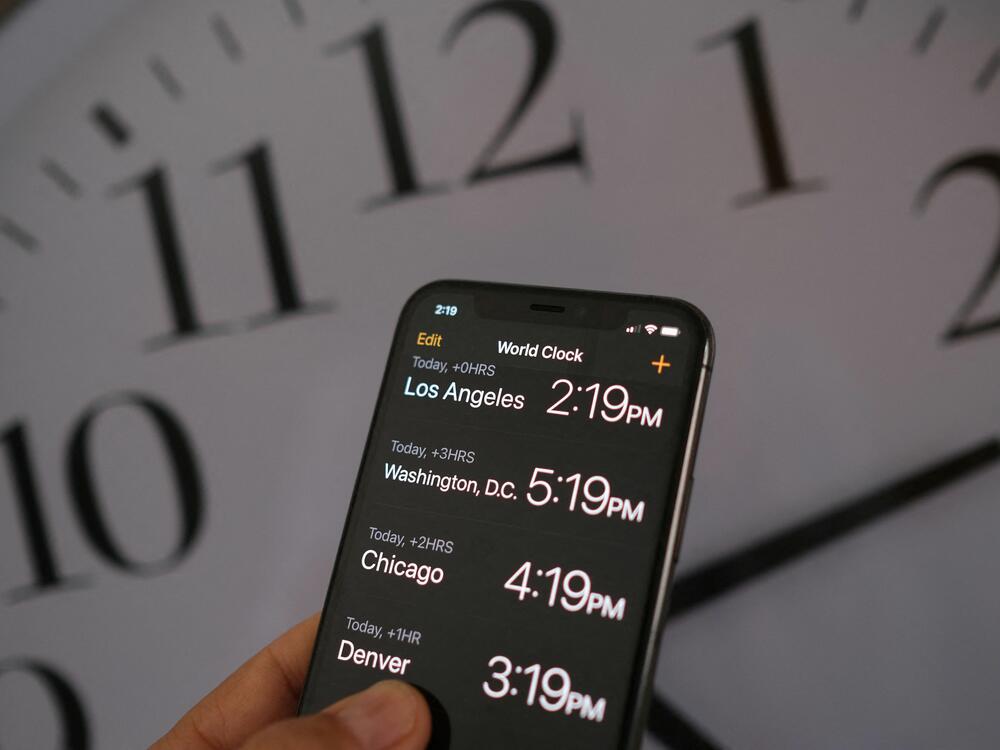 This illustration photo shows a clock in the background of a smartphone showing the time after daylight saving time was implemented in Los Angeles, Calif., on March 15, 2022.