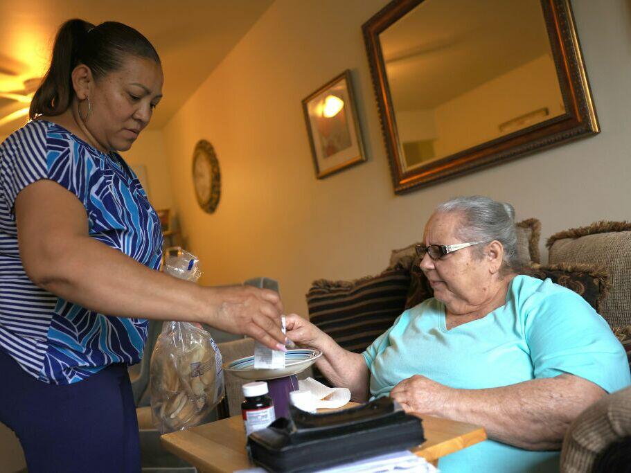 Medicaid pays for nursing home care and for some in-home and community based care, such as aides like Lidia Vilorio, who gives her patient Martina Negron her medicine and a snack in her home on May 5, 2021, in Haverstraw, N.Y.  Proposed cuts to Medicaid could hit such home-based services first, making life harder for family caregivers.
