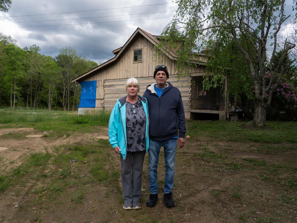 Stuart and Jackie Pacheo stand where the entryway to their home of 30 years was before the floods from Hurricane Helene washed it away. The circles of the tops of foundation pillars in the dirt behind them are all that remain of the Pacheo home.