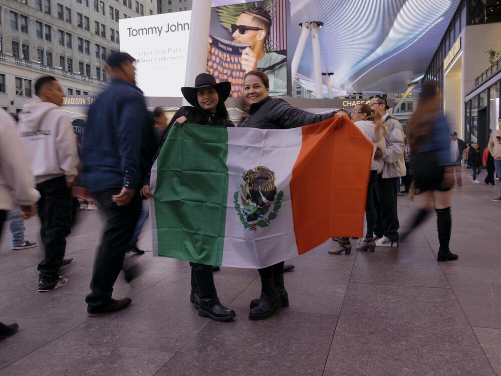 Marcela Rivera (left) and Lidar Portela (right) hold up a Mexican flag and pose for a portrait as fans navigate the bustling block outside of Madison Square Garden before the start of Los Tigres del Norte's show.