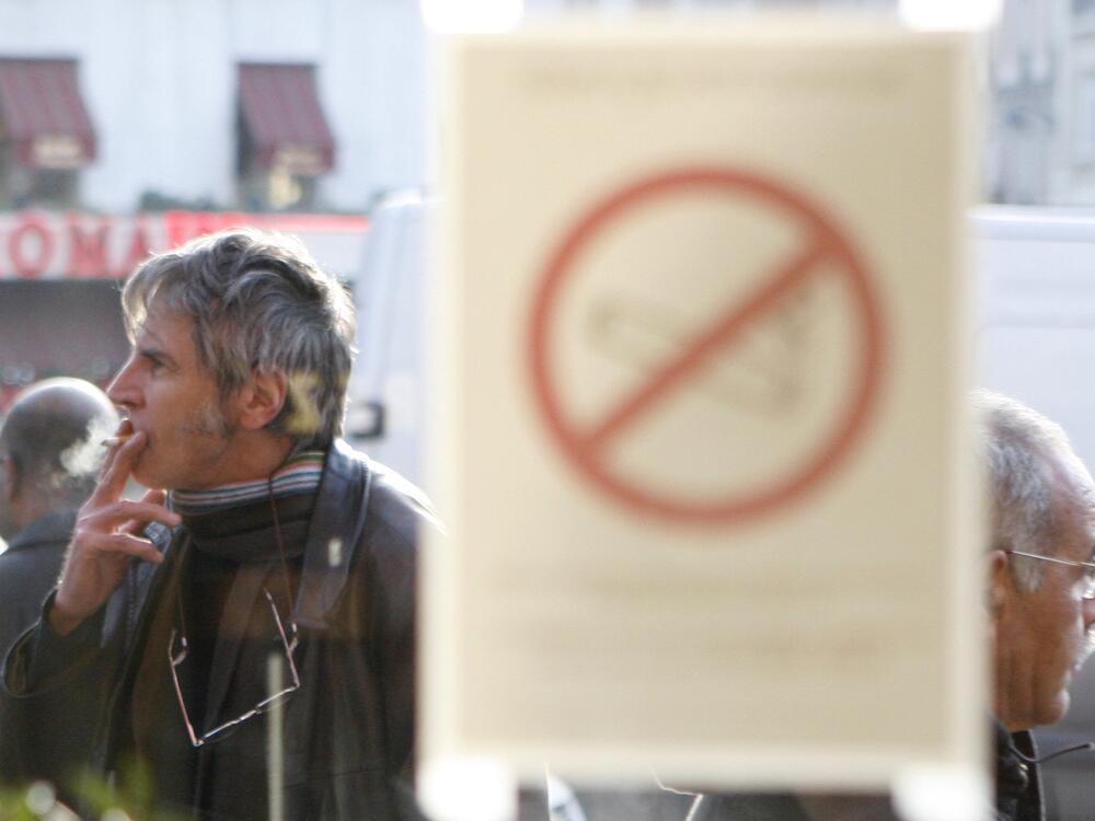 A man smokes a cigarette outside a brasserie, in front of a sticker on a window reading: 