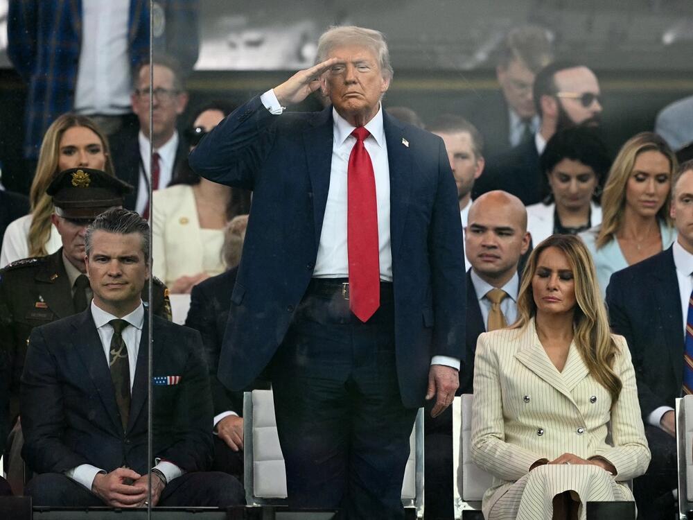 President Trump stands and salutes, flanked by Secretary of Defense Pete Hegseth and First Lady Melania Trump.