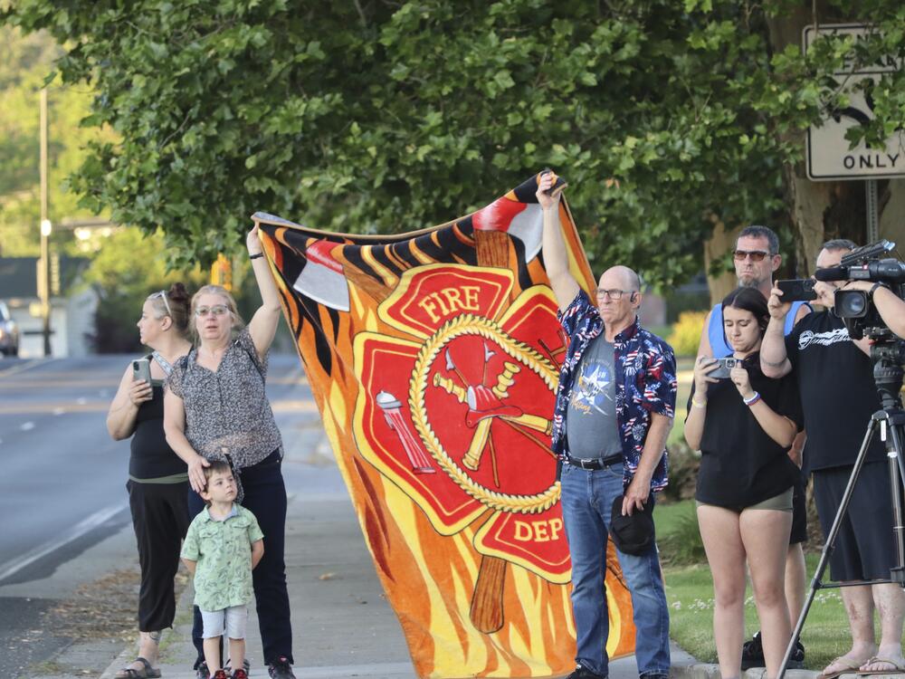 People watch a procession from Kootenai Health headed to Spokane, Wash., after two firefighters were killed Sunday when they were ambushed by sniper fire while responding to a blaze in a northern Idaho mountain community, in Coeur d'Alene, Idaho.