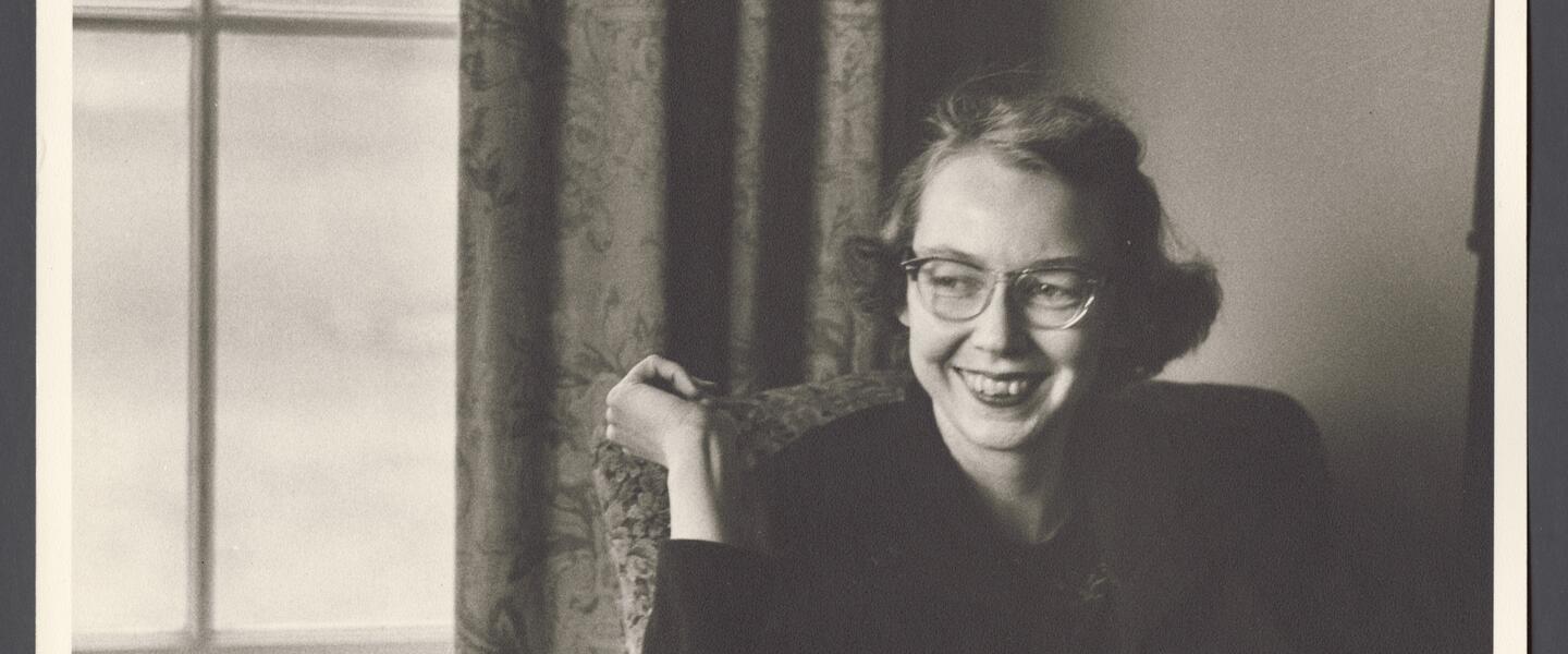 Flannery O’Connor, smiling, sitting by a window.