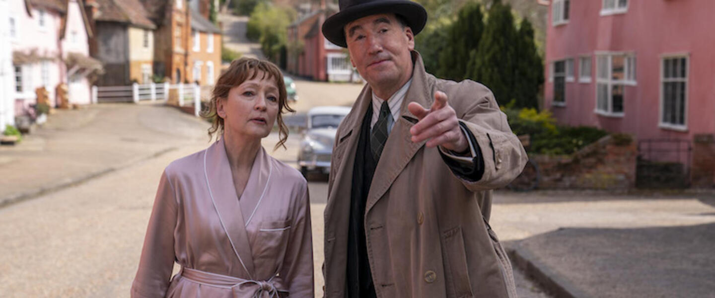 A woman in a robe and a man in a hat and coat looking down a sunny street.