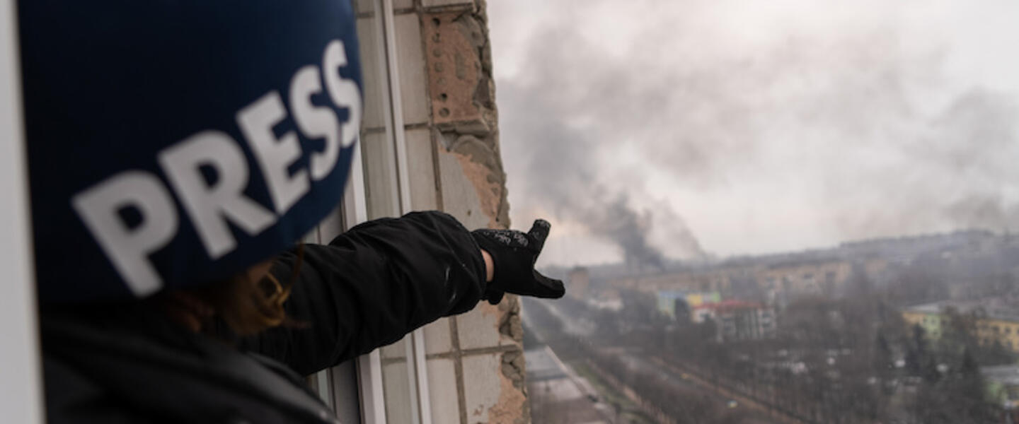 A reporter pointing out a window towards an explosion.