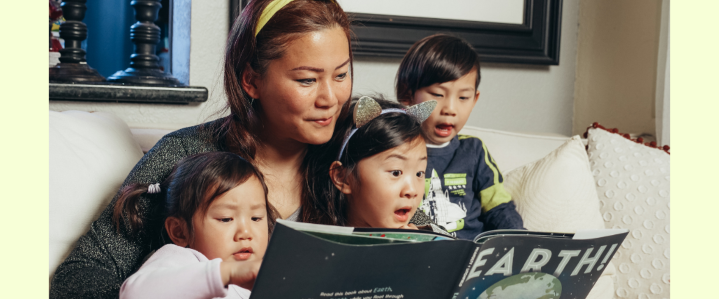 A grownup sits with three kids as they all look at a book.
