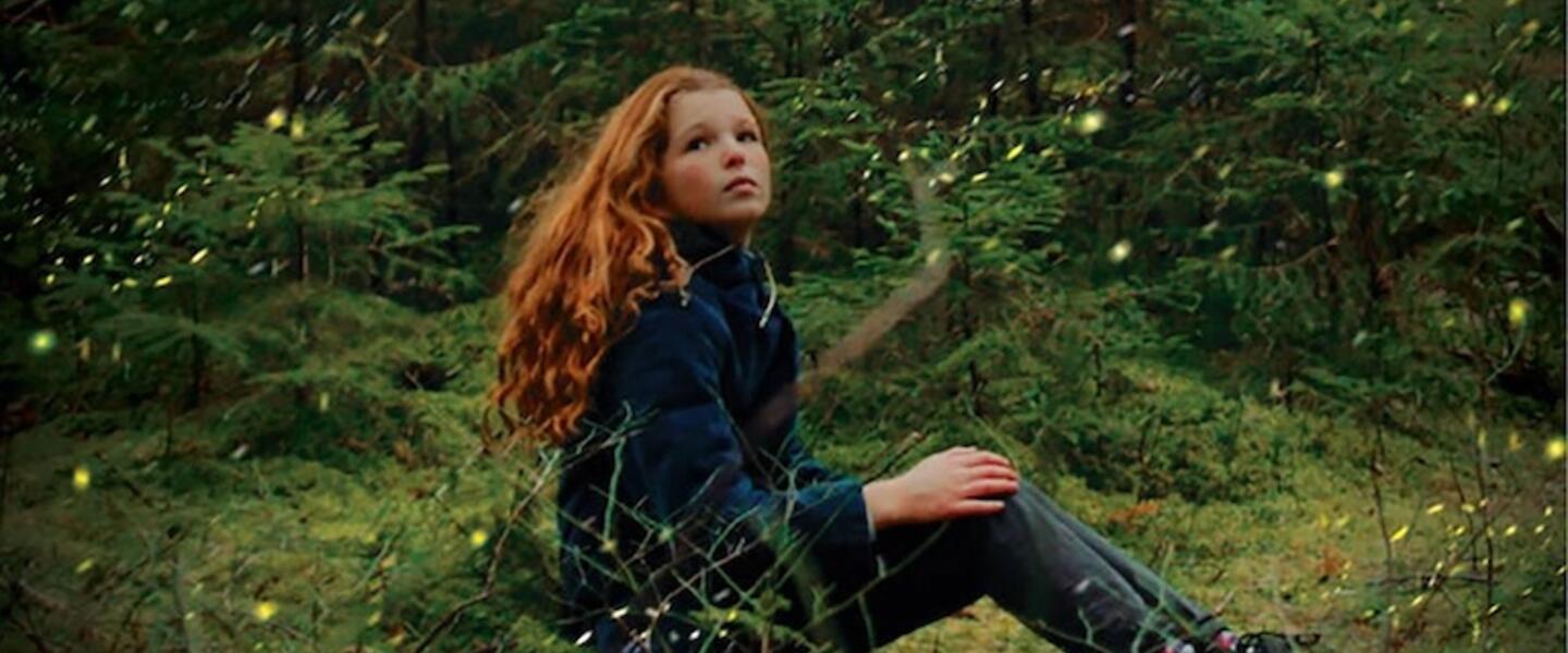 A young girl sitting in a forest surrounded by fireflies
