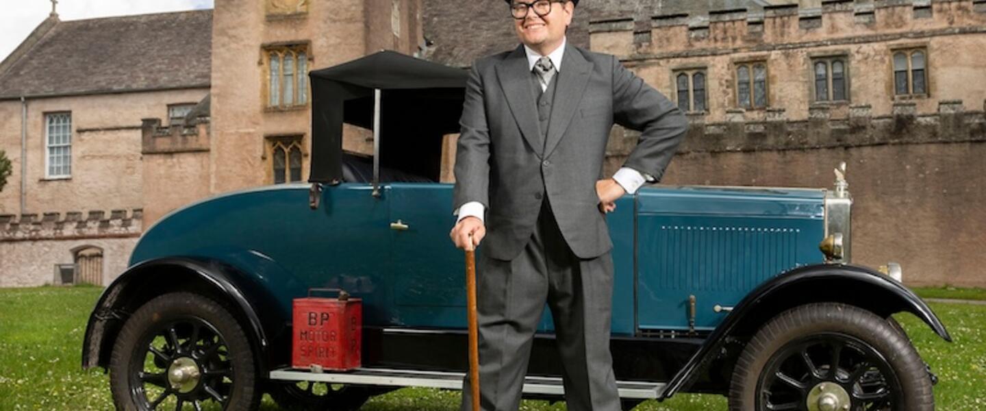 A man in a suit with a cane standing in front of a car from the 20s.