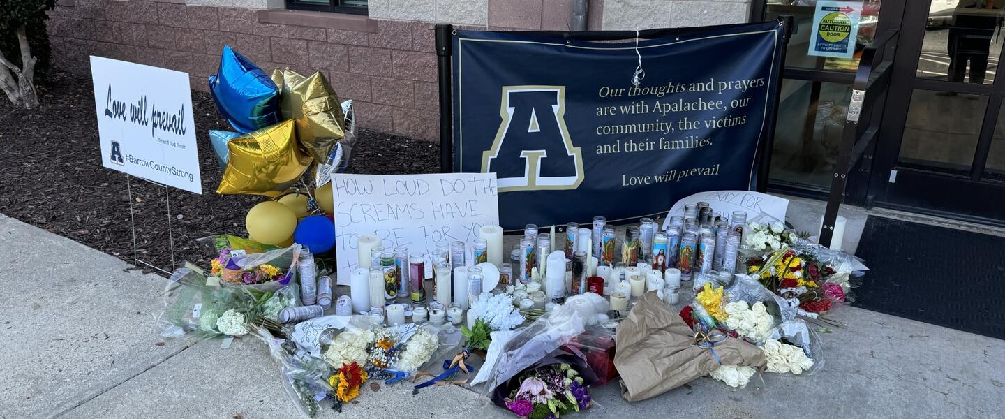 Flowers, balloons, signs, candles and other symbols of support are shown on a sidewalk next to a door.