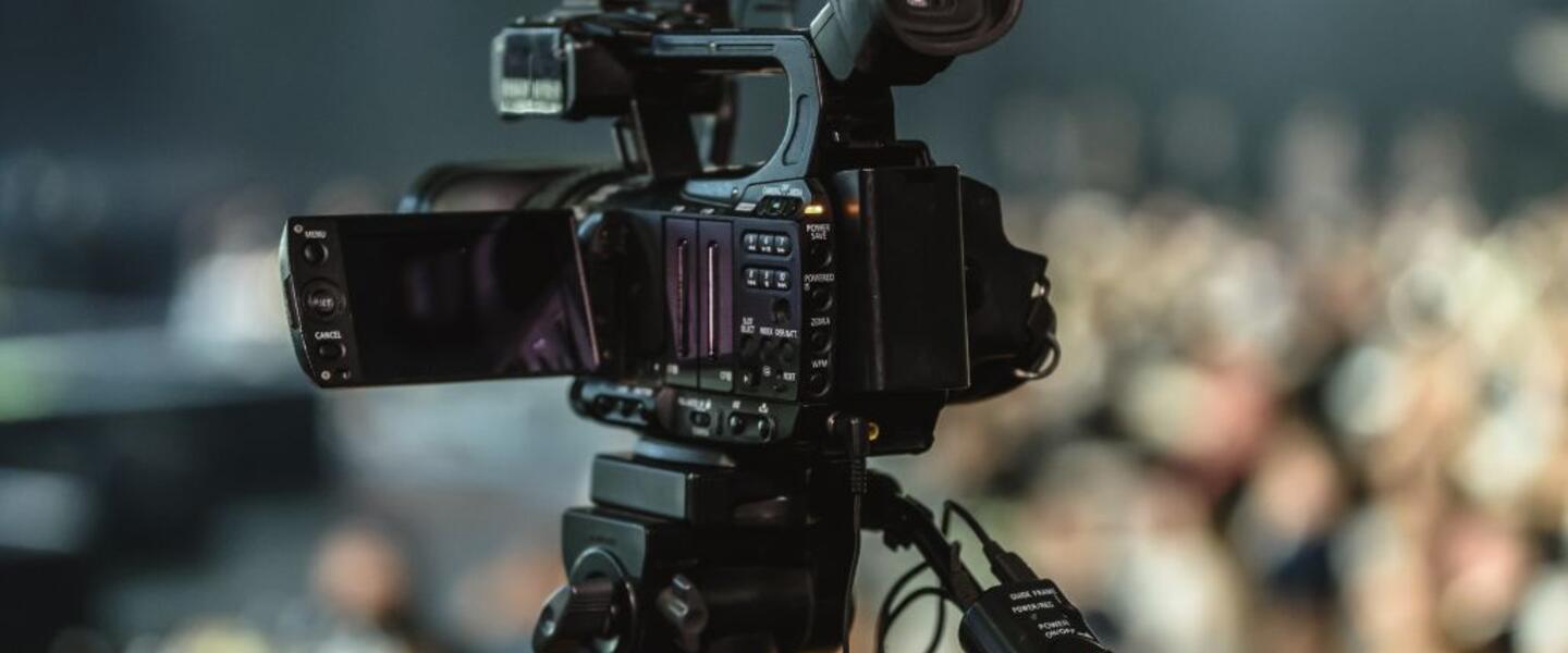lose-up of a video camera mounted on a tripod, with a blurred audience in the background.