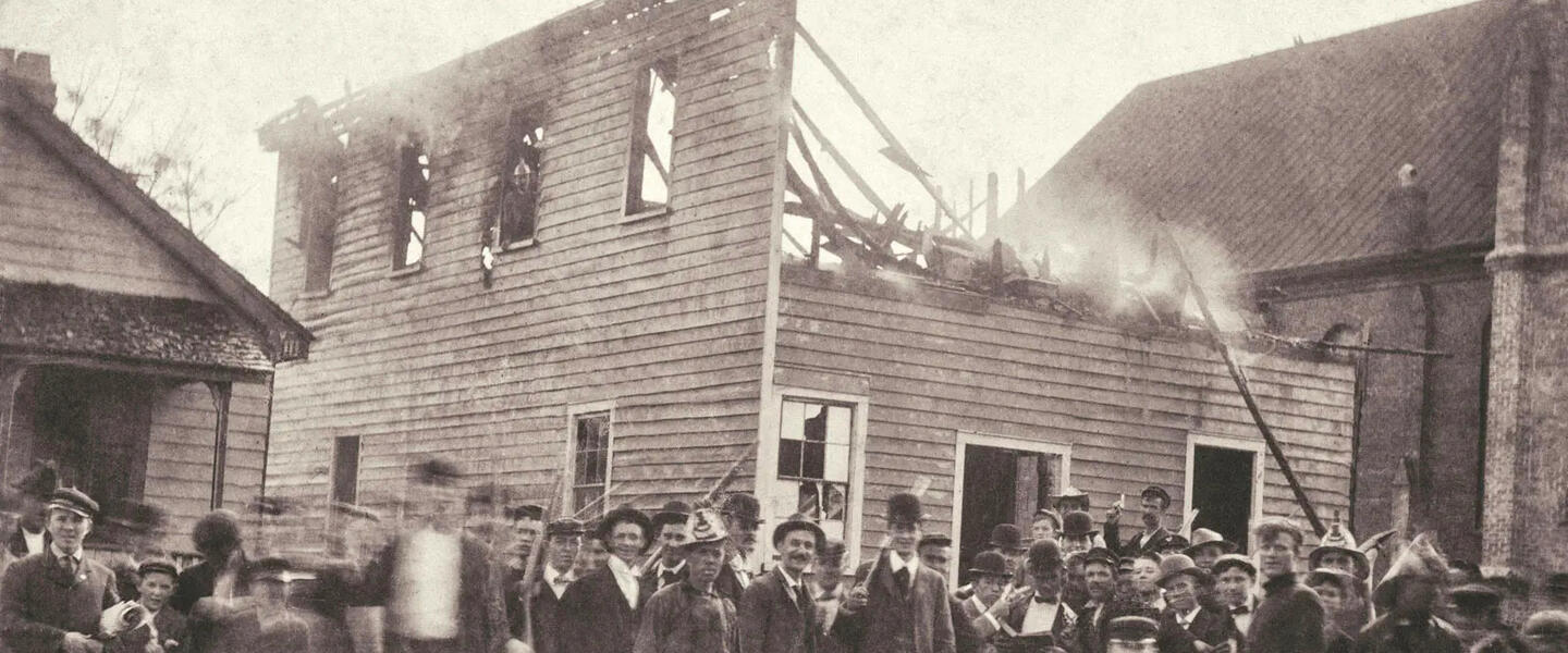 A mob standing in front of a burning building in a sepia photo.