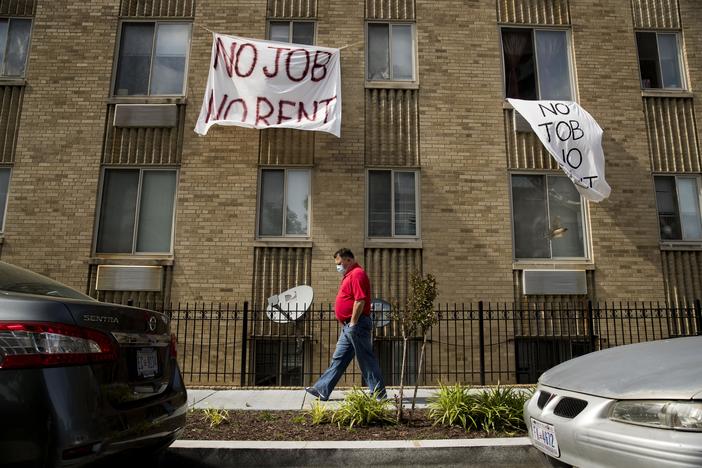 Cloth signs that read "No Job No Rent" hang from the windows of an brown brick apartment building. In front of them, a man wearing jeans and a red shirt and a face mask walks on the sidewalk.