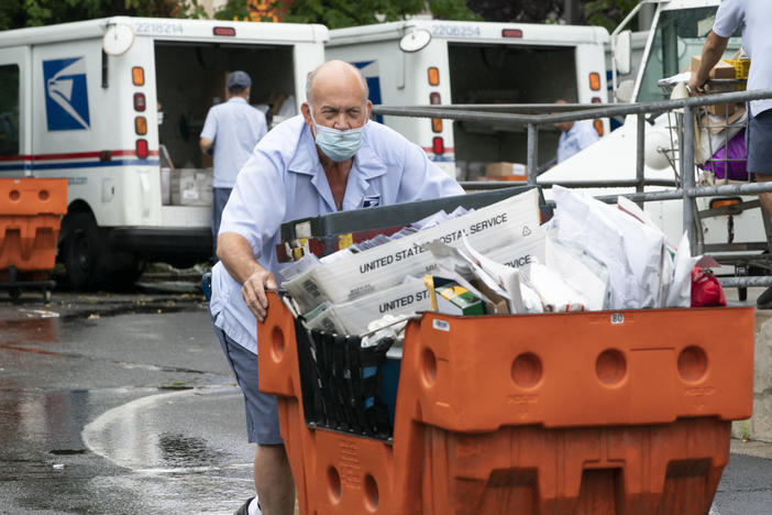 A male U.S. postal worker pushes an orange cart of letter mail towards the camera on a wet day. In the background, two other postal workers unload U.S. Postal Service trucks of mail.