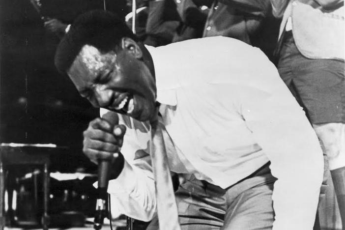 Otis Redding on his knees, singing into a microphone. Behind him, three men play trumpets.